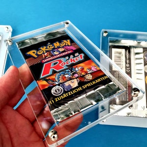 Può includere: Una teca di esposizione in acrilico trasparente con un pacchetto di carte da gioco Pokemon Team Rocket sigillato all'interno. Il pacchetto presenta un design rosso, nero e bianco con le parole "Pokemon Sammelkartenspiel Team Rocket" e "11 Zusätzliche Spielkarten".