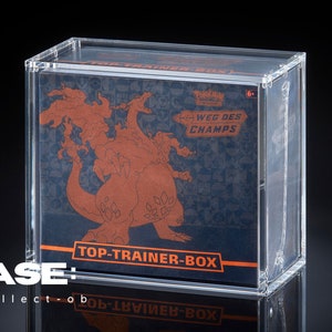 Op de afbeelding: Een heldere acryl display case met een Top Trainer Box. De doos heeft een vurige oranje illustratie van een draakachtig wezen op een donkerblauwe achtergrond. Tekst bevat "WEG DES CHAMPS" en "TOP-TRAINER-BOX".