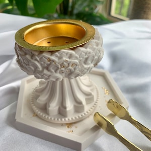May include: White ceramic incense burner with a gold rim and a hexagonal base. The burner has a decorative floral design and is accompanied by gold tongs.