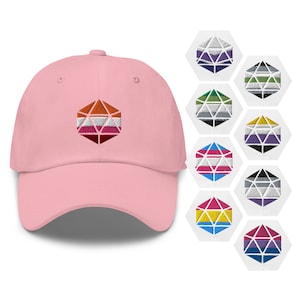 Op de afbeelding: Een roze baseballpet met een veelkleurig geborduurd geometrisch ontwerp. Het ontwerp is een gestileerde weergave van een d20 dobbelsteen met de kleuren van de lesbische pride-vlag.