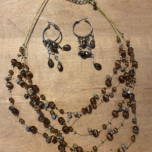 Può includere: Una collana multifilo con perline marroni e dettagli dorati, abbinata a orecchini pendenti coordinati. La collana ha un design a strati con più file di perline.