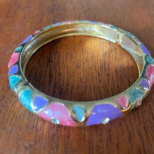 Op de afbeelding: Een gouden armband met een kleurrijk emaille ontwerp. Het emaille is in tinten roze, paars en turquoise. De armband heeft een scharnierende sluiting.