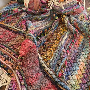 Puede incluir: Una colorida manta de ganchillo texturizada con hilos rosas, azules, verdes y amarillos. La manta tiene un patrón complejo y está colocada sobre una superficie. También se ven varios ovillos de lana y flores.