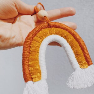 May include: A handmade macrame rainbow wall hanging with a wooden bead at the top. The rainbow is made with orange, yellow, and white yarn and has white tassels.