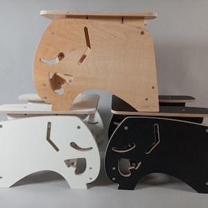Puede incluir: Una pila de cuatro taburetes de madera en acabados natural, blanco y negro. Cada taburete tiene un dise&ntilde;o de recorte &uacute;nico en los lados y una peque&ntilde;a plataforma. Los taburetes est&aacute;n construidos con tornillos visibles y tienen una est&eacute;tica moderna y minimalista.
