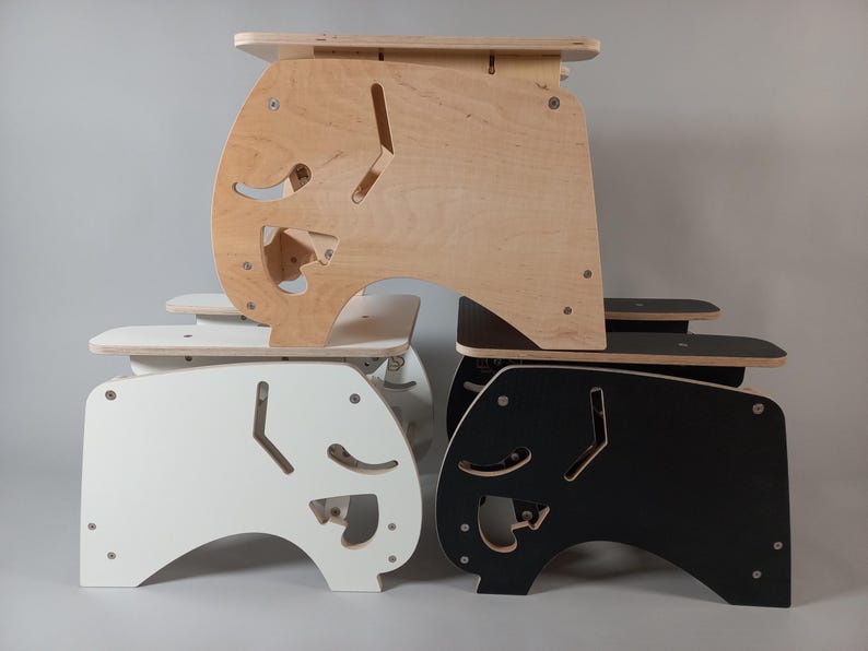 Puede incluir: Una pila de cuatro taburetes de madera en acabados natural, blanco y negro. Cada taburete tiene un dise&ntilde;o de recorte &uacute;nico en los lados y una peque&ntilde;a plataforma. Los taburetes est&aacute;n construidos con tornillos visibles y tienen una est&eacute;tica moderna y minimalista.