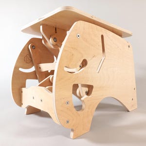 Puede incluir: Un escritorio de pie ajustable de madera de color claro. El escritorio tiene una parte superior rectangular y una base compleja y dise&ntilde;ada con formas recortadas y herrajes visibles. El escritorio est&aacute; dise&ntilde;ado para ser ajustable para uso ergon&oacute;mico.