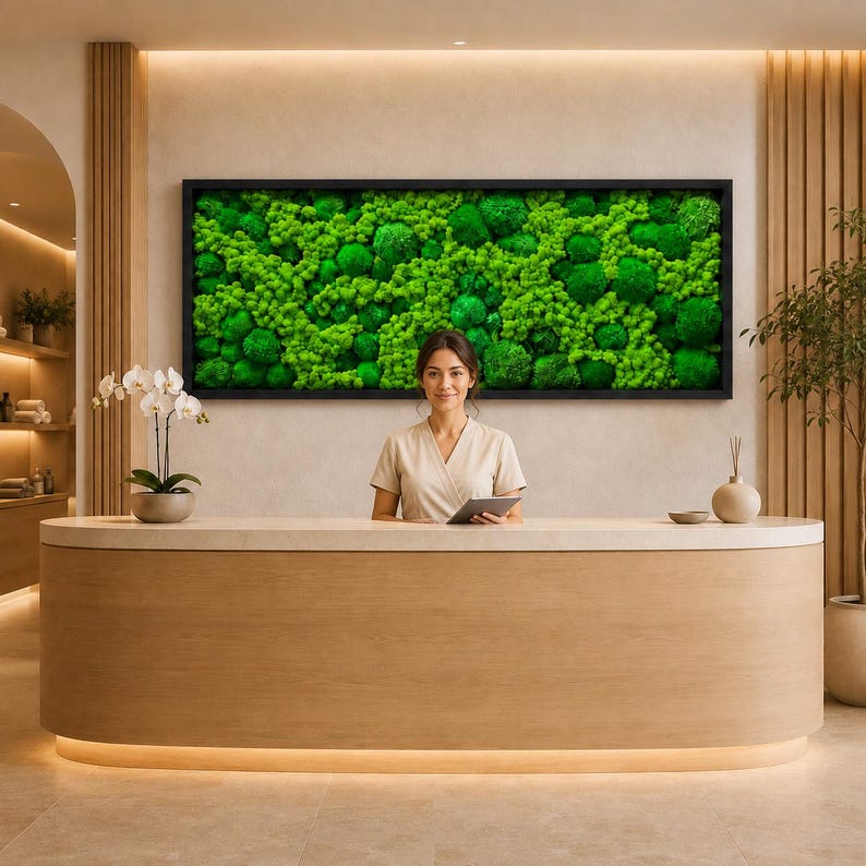 Op de afbeelding: Een lang, rechthoekig wandkunstwerk met een dichte opstelling van levendig groen geconserveerd mos in een zwarte lijst, gemonteerd boven een gebogen, lichtgekleurde houten receptiebalie met een glad oppervlak en ge&iuml;ntegreerde verlichting.