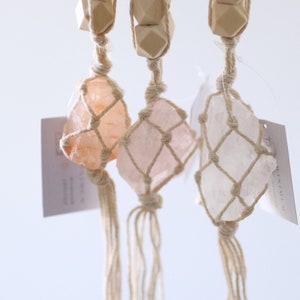 Puede incluir: Tres adornos colgantes de cristal. Cada adorno presenta un cristal en bruto envuelto en una red de macramé, suspendido de un cordón de fibra natural. Cuentas de madera se colocan encima de los cristales. Los cristales son de varios tonos de rosa y melocotón.