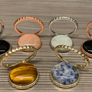 May include: Six different phone ring holders with various gemstone inlays. The rings are made of silver, gold, rose gold, and black metal. The gemstones include black onyx, tiger's eye, blue sodalite, white agate, and pink quartz.