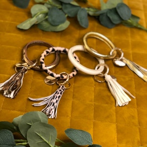 Può includere: Quattro portachiavi con cinturini di diversi colori e motivi. I cinturini sono in pelle e hanno un anello di metallo dorato. Ogni portachiavi ha un fiocco attaccato all'anello. I fiocchi sono fatti di diversi materiali, tra cui pelle, pelliccia e metallo.