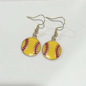 May include: A pair of softball earrings. Each earring features a yellow softball design with red stitching and gold-colored ear hooks. The earrings are designed to dangle.
