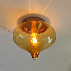 Puede incluir: Una lámpara de techo vintage con una pantalla de cristal ámbar en forma de lágrima. La bombilla es visible dentro del cristal, y la lámpara tiene una base de metal color latón. La luz está encendida.