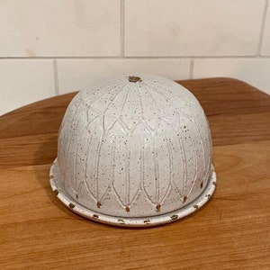 May include: A white ceramic butter dish with a domed lid and a matching base. The dish has a speckled texture and a decorative pattern of vertical lines and small gold accents. The butter dish is sitting on a wooden surface.