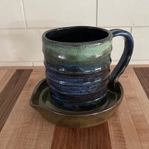 May include: A ceramic mug with a ribbed texture and a handle, featuring a gradient glaze that transitions from a deep blue at the base to a lighter green at the rim. The mug sits inside a matching circular ceramic saucer with a dark brown glaze.