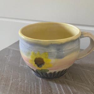 May include: A ceramic mug featuring a hand-painted sunflower design against a soft pastel background of yellow, blue, and pink hues. The base of the mug is decorated with a dark, scalloped pattern, and it includes a single side handle.