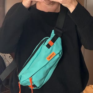 May include: A teal bum bag with black straps and orange detailing. The bag has a rectangular shape with a zip closure and a small orange tag with text. The bag is worn across the body.