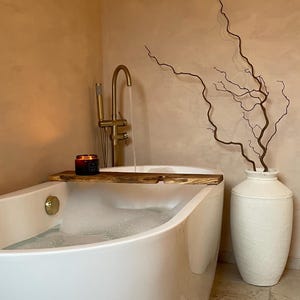 May include: A contemporary bathroom with a white bathtub filled with bubbles. A wooden bath tray holds a candle. A gold tap pours water. A white vase with branches sits beside the tub. The walls are painted a warm peach colour.