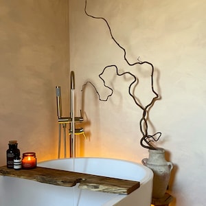 May include: A white bathtub with a wooden bath tray, two small bottles, and a candle. The bathtub is in a bathroom with a light brown wall and a vase with a dried branch.