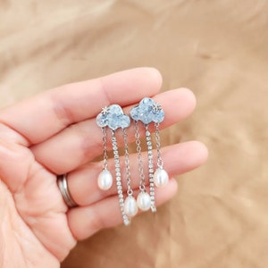Peut inclure: Boucles d'oreilles pendantes argentées avec un motif nuage et un détail étoile. Les boucles d'oreilles ont trois gouttes de perles suspendues à chaque nuage.