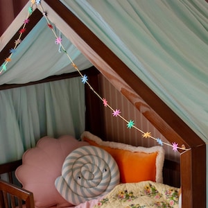Pode incluir: Uma estrutura de cama de madeira escura em forma de casa, coberta com tecido azul claro. A cama inclui uma manta acolchoada com flores amarelas, uma almofada rosa em forma de concha, uma almofada redonda com espirais e uma almofada laranja, com guirlanda.