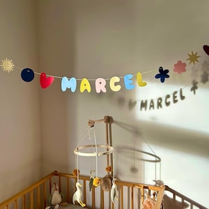 May include: A nursery scene featuring a wooden crib with a mobile and a banner that spells out "MARCEL" in colorful letters. The banner includes shapes like hearts, flowers, and stars. Soft toys are visible inside the crib.