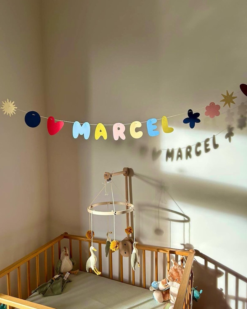 May include: A nursery scene featuring a wooden crib with a mobile and a banner that spells out "MARCEL" in colorful letters. The banner includes shapes like hearts, flowers, and stars. Soft toys are visible inside the crib.