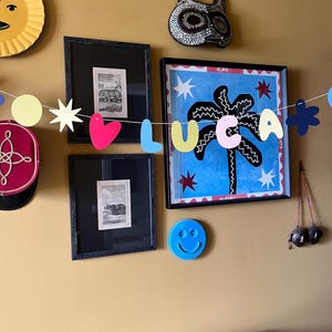 May include: A collection of wall art and decorations on a mustard-colored wall. Includes framed prints, a palm tree painting, a smiley face, and a garland spelling "LUCA". A sun-shaped decoration and a decorative mask are also visible.