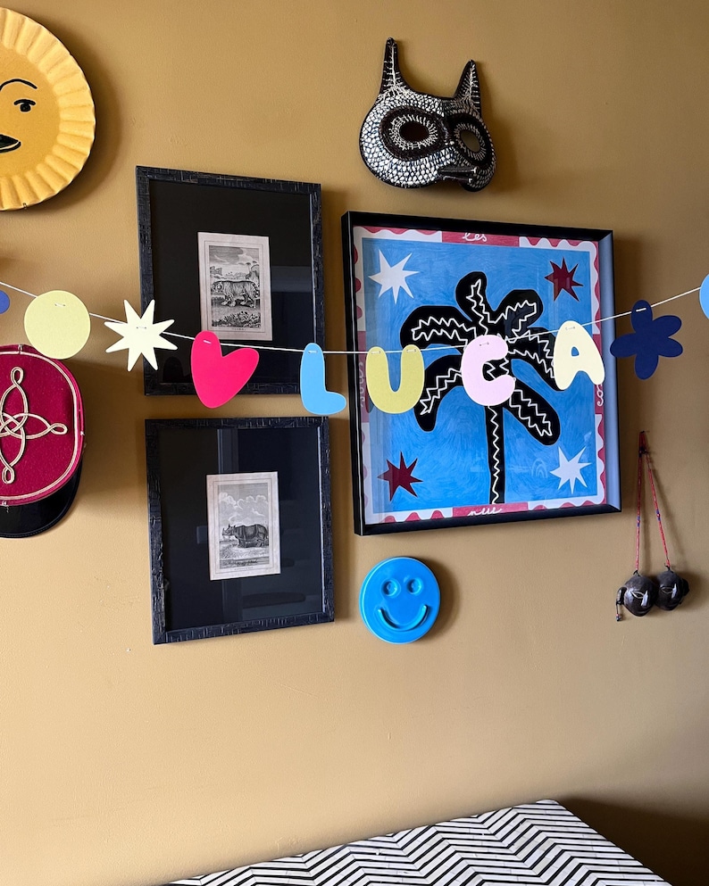 May include: A collection of wall art and decorations on a mustard-colored wall. Includes framed prints, a palm tree painting, a smiley face, and a garland spelling "LUCA". A sun-shaped decoration and a decorative mask are also visible.