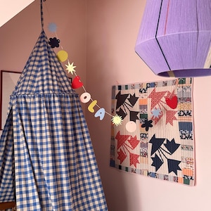 May include: A blue and white checkered canopy hangs in a pink room, with a garland that spells "OLA". A colorful quilt with star and heart shapes hangs on the wall, and a purple lampshade is suspended from the ceiling.