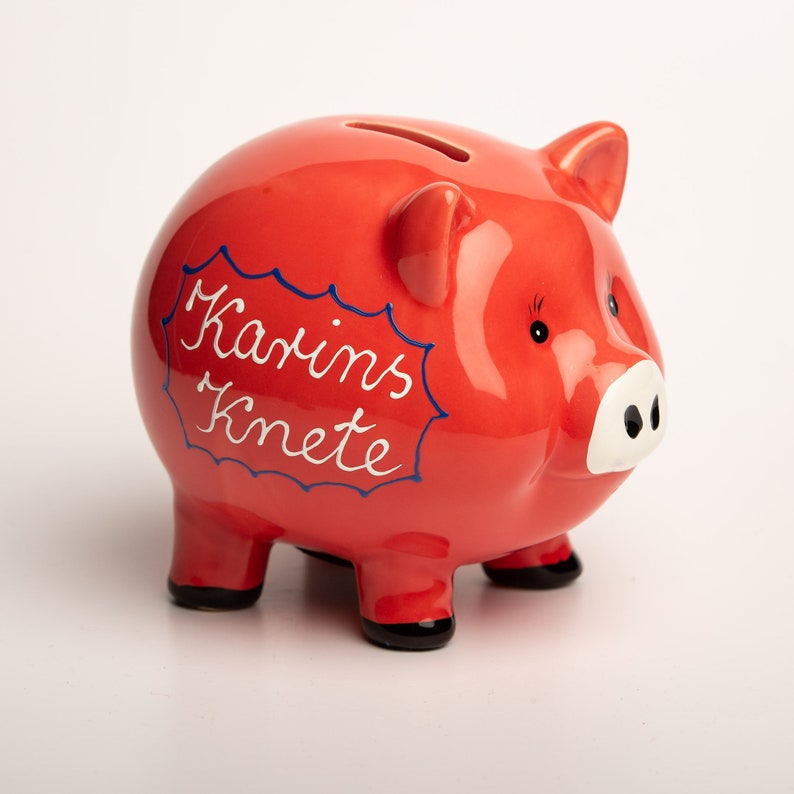 May include: A red ceramic piggy bank with a white and blue inscription that reads "Karins Knete".