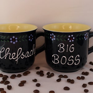 May include: Two blue ceramic mugs with yellow rims and white lettering. The mug on the left says "chefsache" and the mug on the right says "BIG BOSS". Both mugs have a floral design around the top.