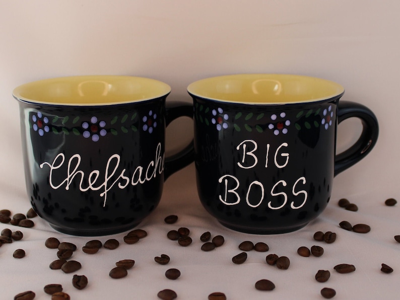 May include: Two blue ceramic mugs with yellow rims and white lettering. The mug on the left says "chefsache" and the mug on the right says "BIG BOSS". Both mugs have a floral design around the top.