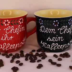 May include: Two ceramic coffee mugs with a folk art design. The red mug says "Der Chefin ihr Kaffeepott" and the blue mug says "Dem Chef sein Kaffeepott".