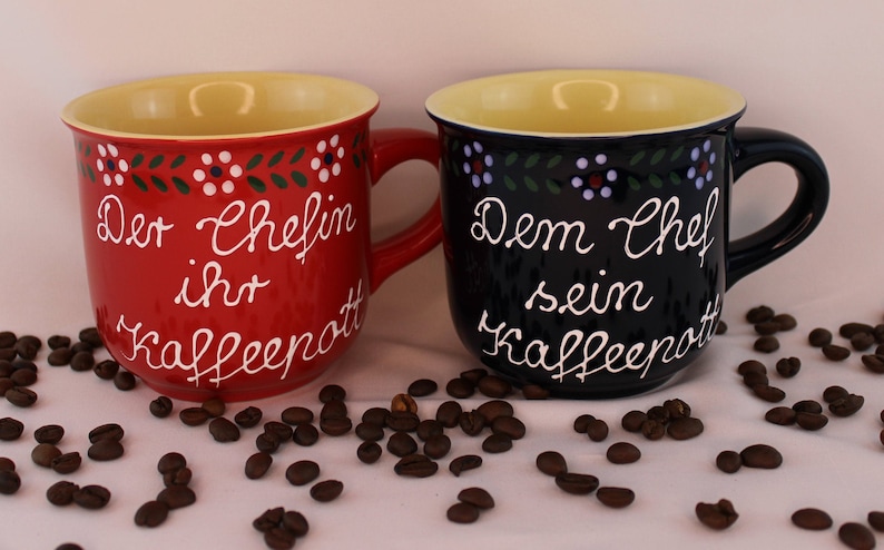 May include: Two ceramic coffee mugs with a folk art design. The red mug says "Der Chefin ihr Kaffeepott" and the blue mug says "Dem Chef sein Kaffeepott".