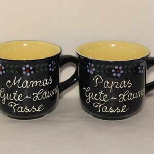 May include: Two black ceramic mugs with yellow interiors and a floral design. One mug says "Mamas Gute-Laune Tasse" and the other says "Papas Gute-Laune Tasse".