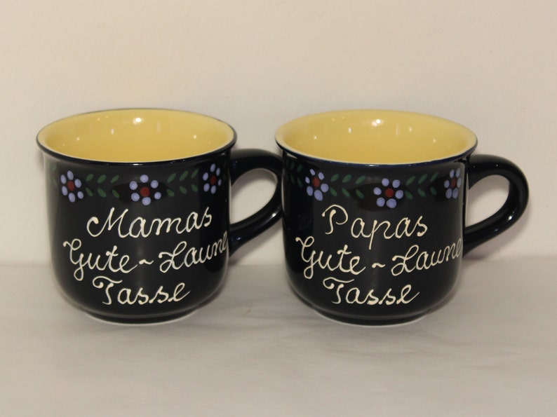 May include: Two black ceramic mugs with yellow interiors and a floral design. One mug says "Mamas Gute-Laune Tasse" and the other says "Papas Gute-Laune Tasse".