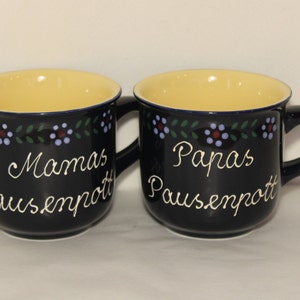 May include: Two blue ceramic mugs with yellow interiors and white lettering. The mugs have a floral design around the top. One mug says "Mamas Pausenpott" and the other says "Papas Pausenpott".