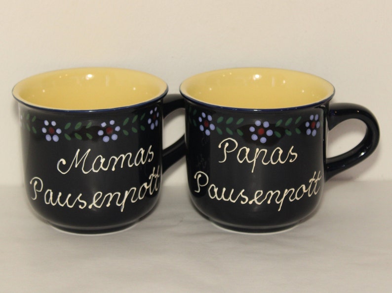 May include: Two blue ceramic mugs with yellow interiors and white lettering. The mugs have a floral design around the top. One mug says "Mamas Pausenpott" and the other says "Papas Pausenpott".