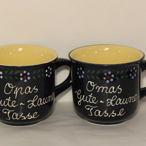 May include: Two black ceramic mugs with yellow interiors and white lettering. The mugs have a floral design around the top and say "Opa's Gute Laune Tasse" and "Oma's Gute Laune Tasse".