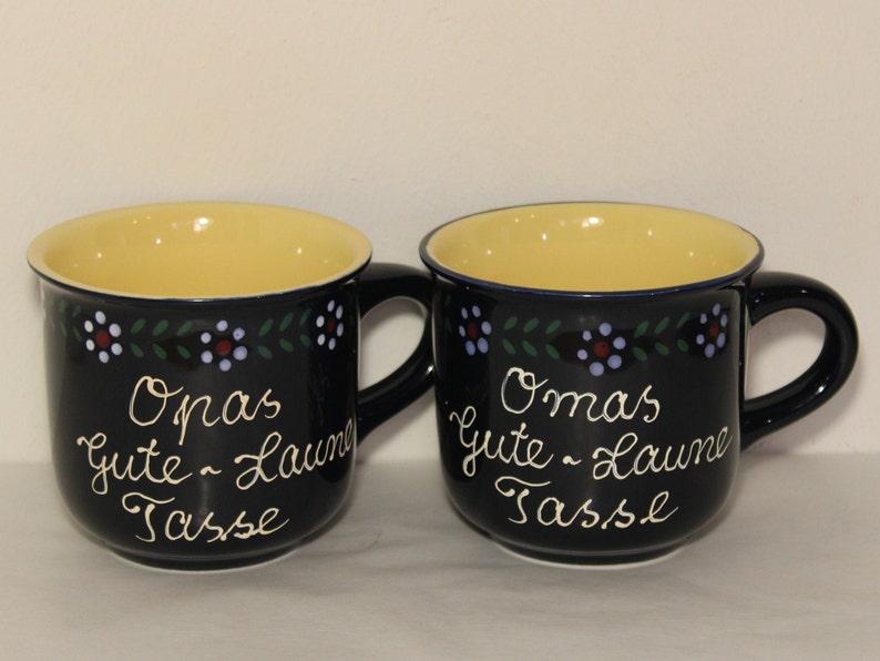 May include: Two black ceramic mugs with yellow interiors and white lettering. The mugs have a floral design around the top and say "Opa's Gute Laune Tasse" and "Oma's Gute Laune Tasse".