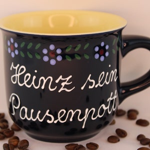 May include: A black ceramic mug with a yellow interior and a white inscription that reads "Heinz sein Pausenpott". The mug is decorated with a blue, green, and red floral pattern around the rim.