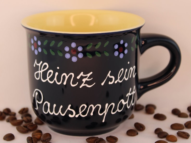 May include: A black ceramic mug with a yellow interior and a white inscription that reads "Heinz sein Pausenpott". The mug is decorated with a blue, green, and red floral pattern around the rim.