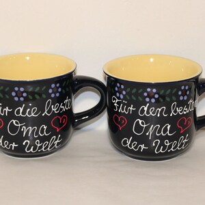 May include: Two blue ceramic mugs with yellow interiors and white lettering. The mugs have a floral design around the top and say "F&uuml;r die beste Oma der Welt" and "F&uuml;r den besten Opa der Welt" with red hearts.