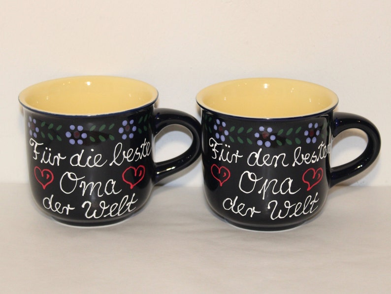 May include: Two blue ceramic mugs with yellow interiors and white lettering. The mugs have a floral design around the top and say "F&uuml;r die beste Oma der Welt" and "F&uuml;r den besten Opa der Welt" with red hearts.