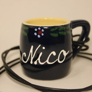 Könnte beinhalten: Eine kleine blaue Keramiktasse mit einem weißen Henkel und einer weißen Aufschrift "Nico". Die Tasse hat ein weißes und grünes Blumenmuster um den oberen Rand. Die Tasse hängt an einer schwarzen Schnur.