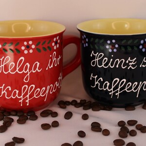 May include: Two ceramic coffee mugs with yellow rims and painted floral designs. The red mug says "Helga ihr Kaffeepott" and the blue mug says "Heinz sein Kaffeepott".