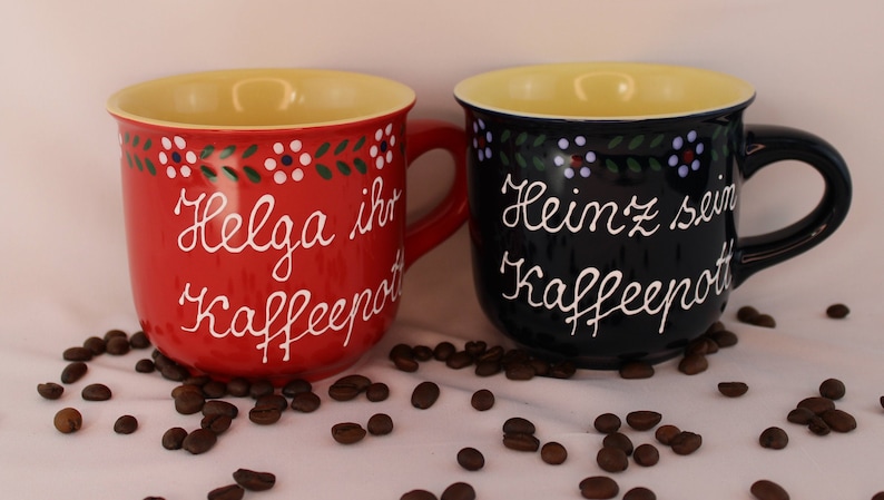 May include: Two ceramic coffee mugs with yellow rims and painted floral designs. The red mug says "Helga ihr Kaffeepott" and the blue mug says "Heinz sein Kaffeepott".