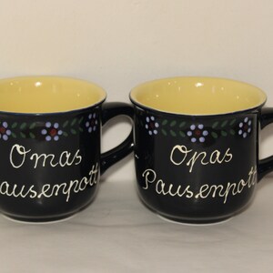 May include: Two black ceramic mugs with yellow interiors and blue and green floral designs. The mugs have white text that reads "Omas Pausenpott" and "Opas Pausenpott".