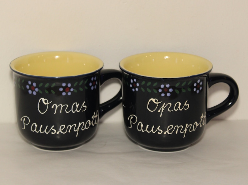 May include: Two black ceramic mugs with yellow interiors and blue and green floral designs. The mugs have white text that reads "Omas Pausenpott" and "Opas Pausenpott".